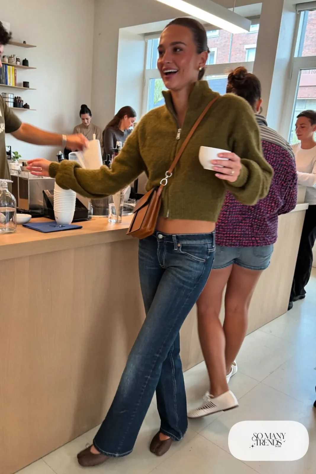 Woman in a short olive zip knit sweater, low-rise dark denim flare jeans and brown leather ballet flats, look 6