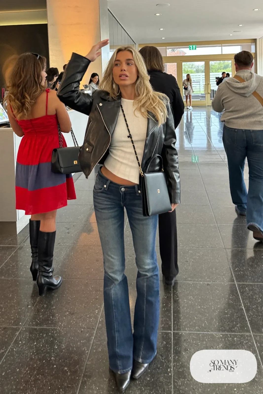Woman in a short black leather jacket, fitted ivory knit top, low-rise indigo flare jeans and black pointed boots, look 15