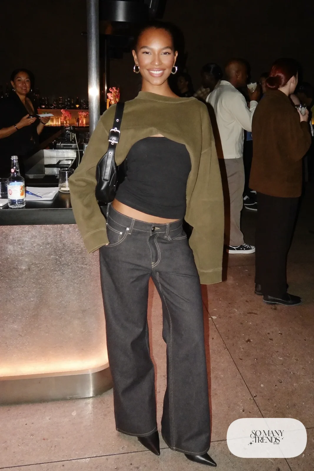 Woman in a sheer olive mesh long-sleeve over a black tank, low-rise dark flare jeans and black pointed boots, look 17