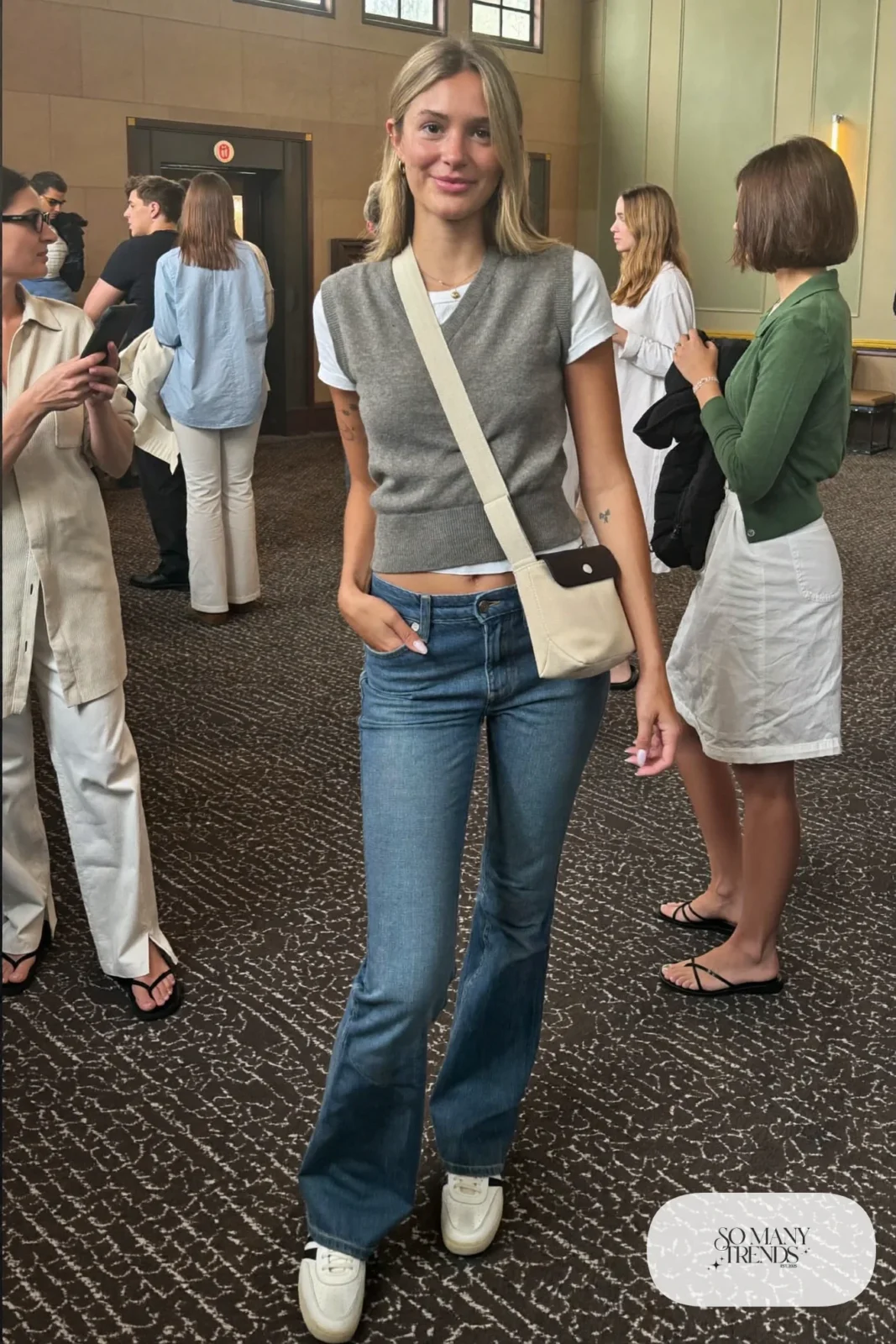 Woman in a gray knit sweater vest, fitted white tee, low-rise mid-blue flare jeans and slim white leather sneakers, look 18