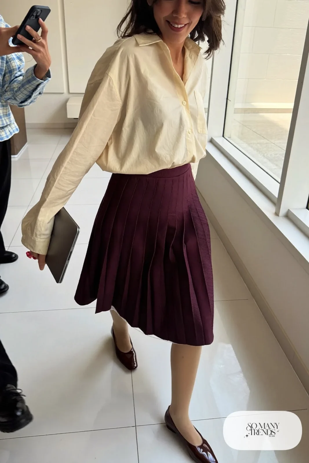 Woman in a plum pleated midi skirt, cream button-up shirt and brown flats in a bright office corridor, outfit 6