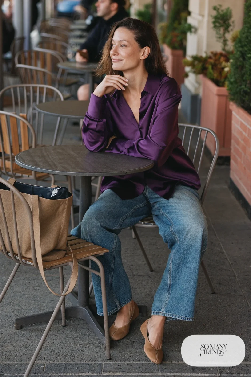 Woman in an eggplant satin shirt, wide-leg medium wash jeans and tan flats at an outdoor cafe, outfit 11