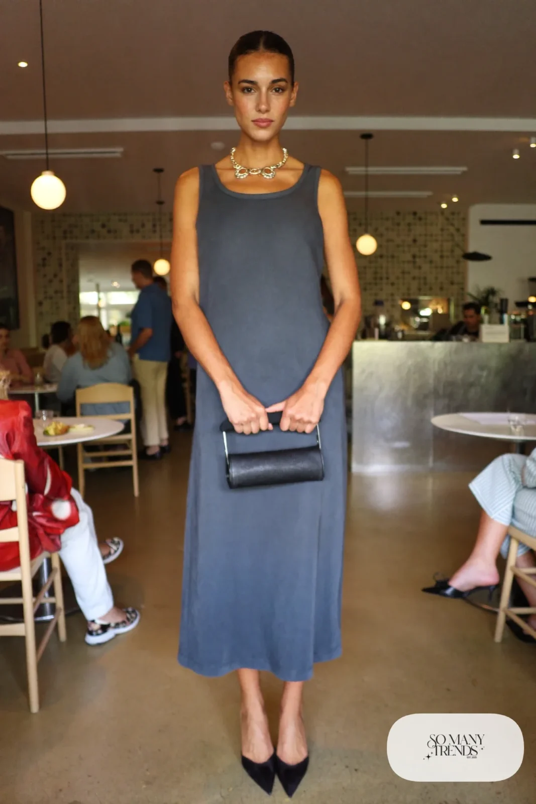 Woman in a slate blue sleeveless column dress with a chunky gold collar necklace, bare legs and black pointed pumps, outfit 1
