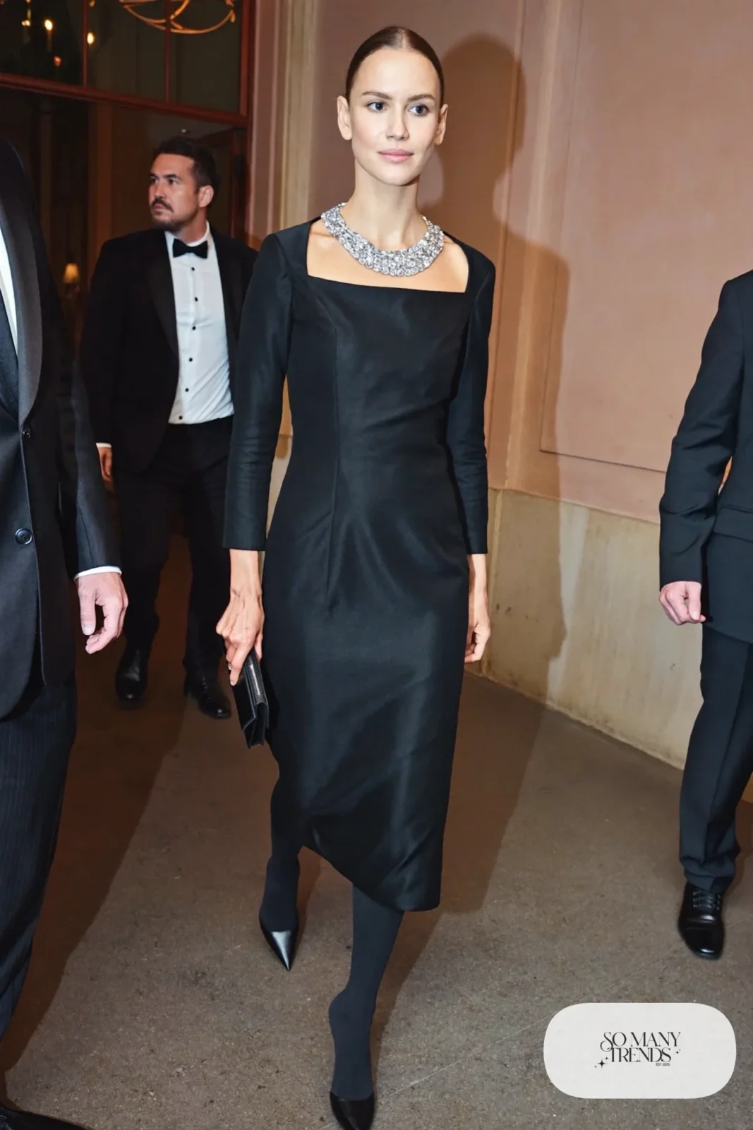Woman in a black square neck dress with a crystal bib necklace, black tights and pointed pumps, outfit 4