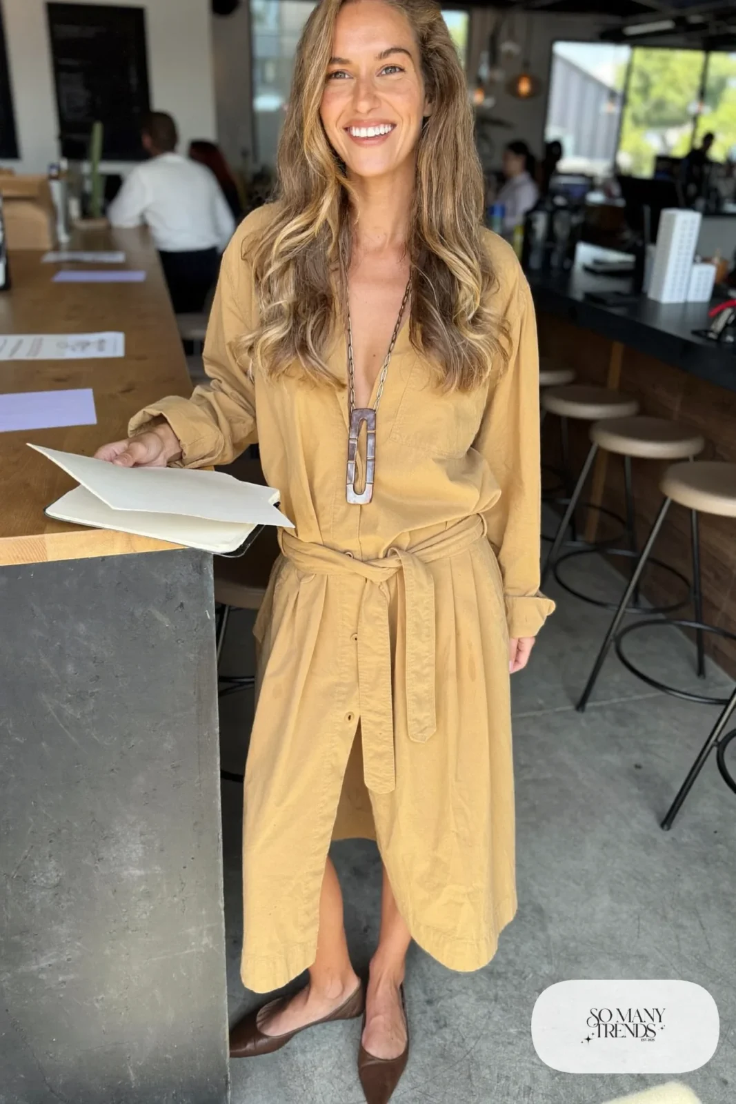 Woman in a camel belted shirt dress with a long pendant necklace and pointed brown flats, outfit 7