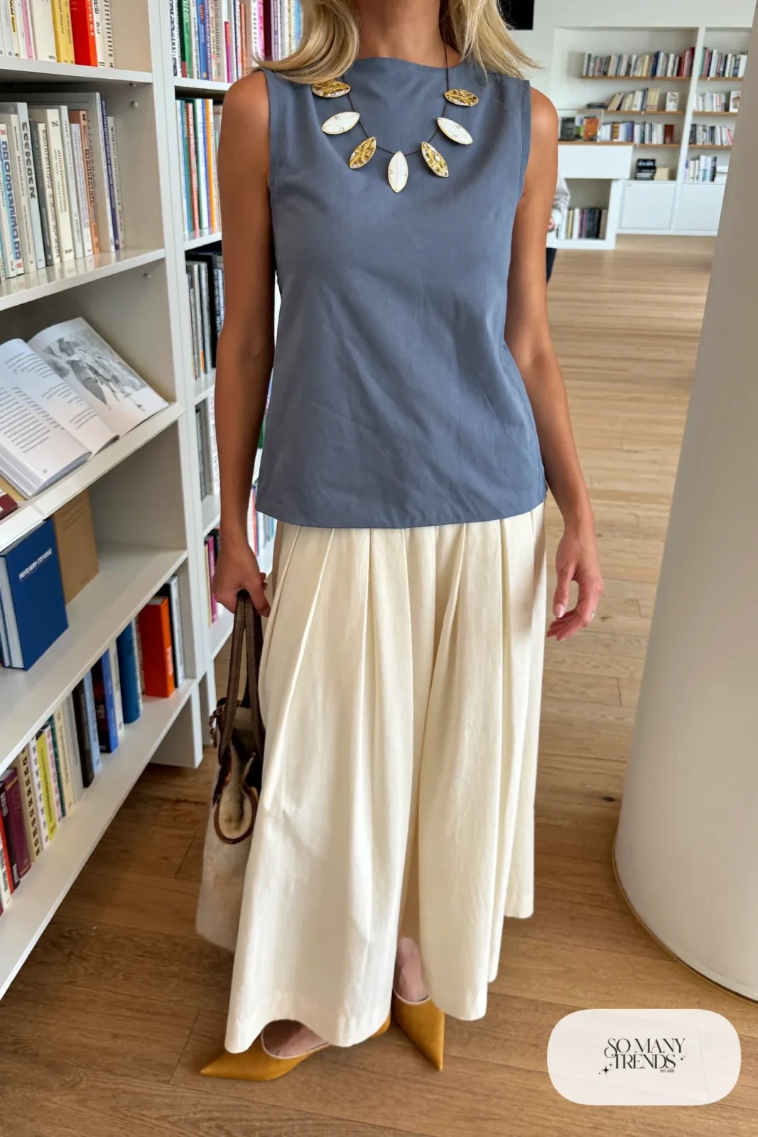 Woman in a slate blue sleeveless shell top with a long cream pleated skirt, a bold enamel necklace and tan flats, outfit 9