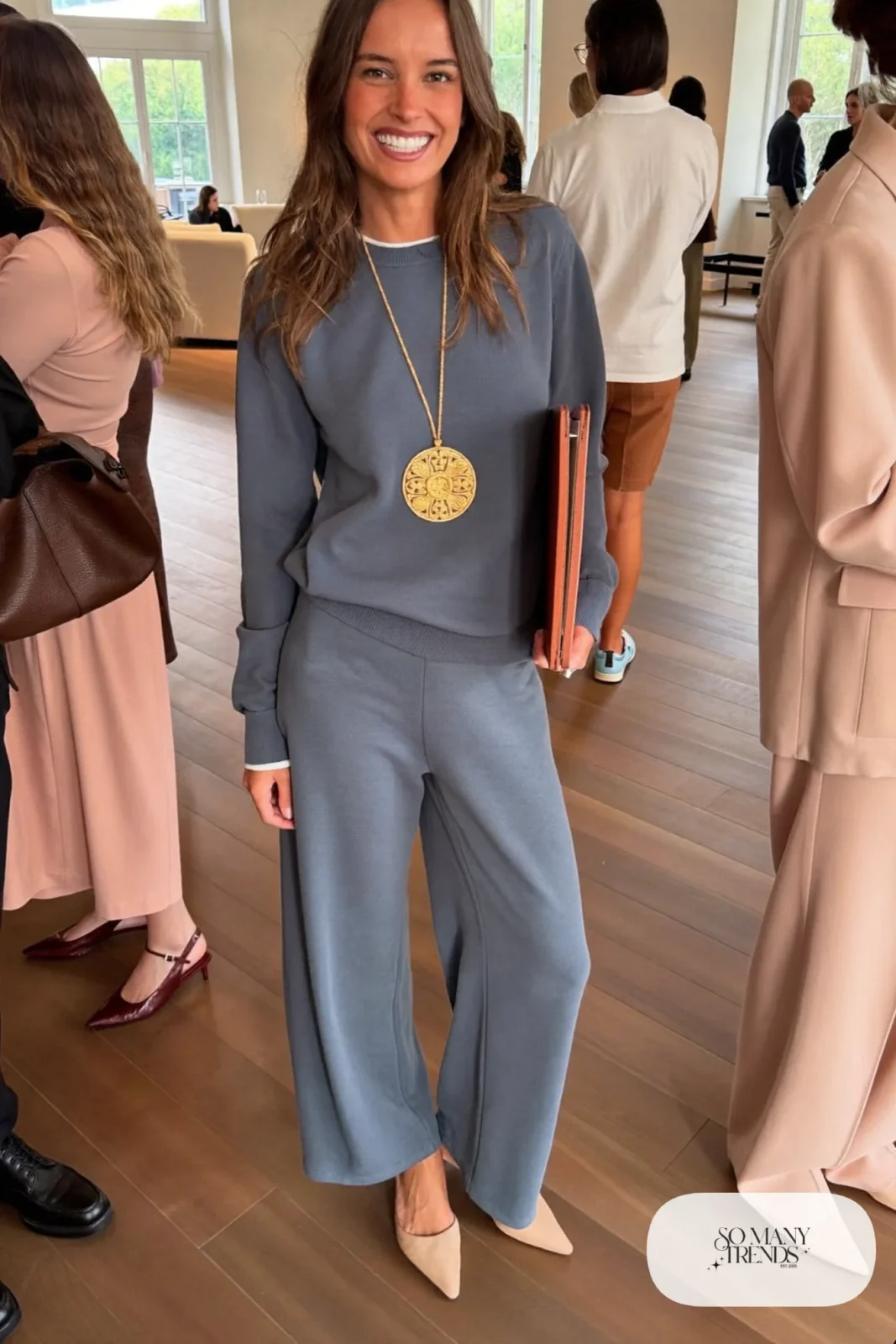 Woman in a slate blue knit top and wide-leg pants set with a large gold medallion necklace and pointed flats, outfit 14