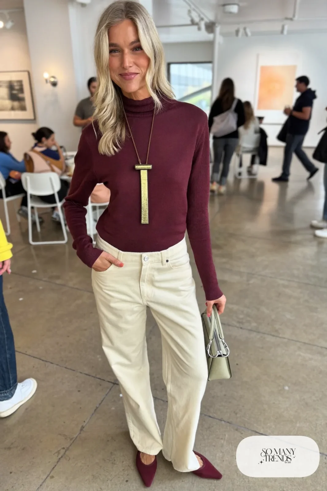 Woman in a wine mock neck top with a long vertical bar necklace, cream straight pants and wine flats, outfit 15