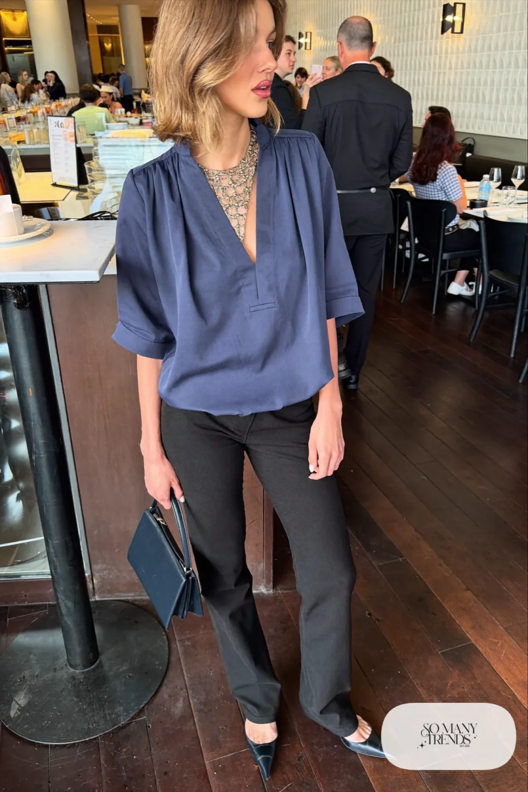 Woman in a slate blue crepe V-neck blouse, slim black pants, bold pendant necklace and black heels, outfit 21