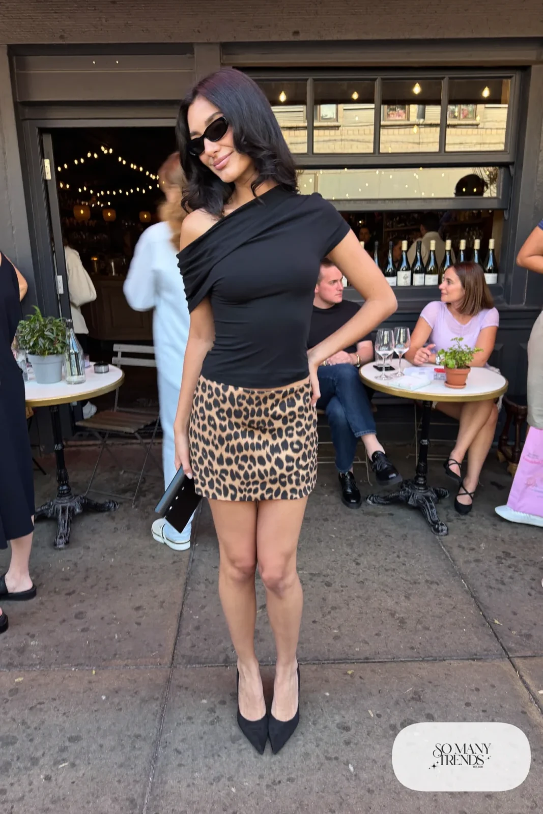 Woman in a black off-shoulder top, leopard print mini skirt, bare legs and black heeled sandals-style closed pumps, outfit 12