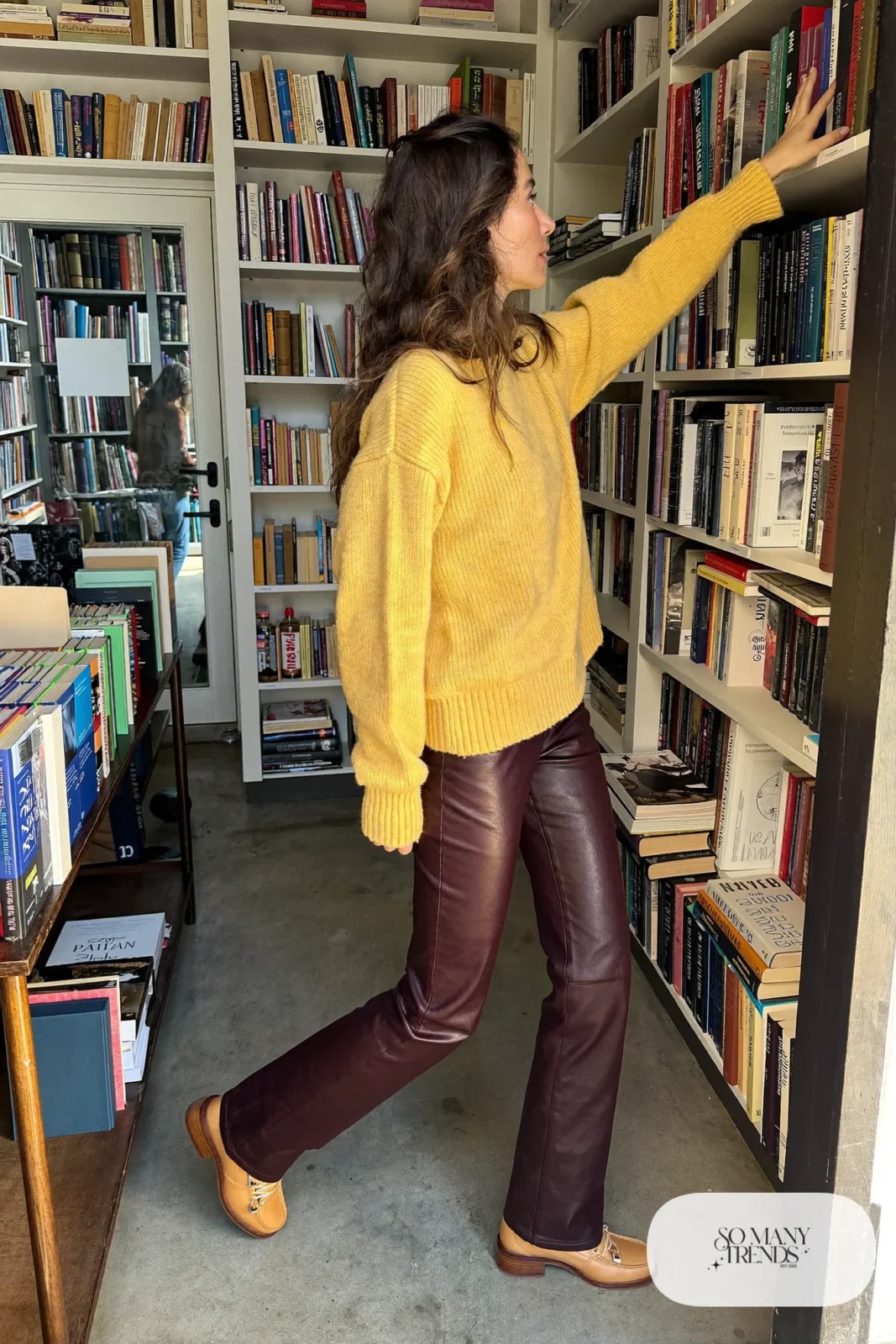 Woman in aubergine straight leather pants, camel oversized sweater and tan loafers in a bookstore, outfit 3