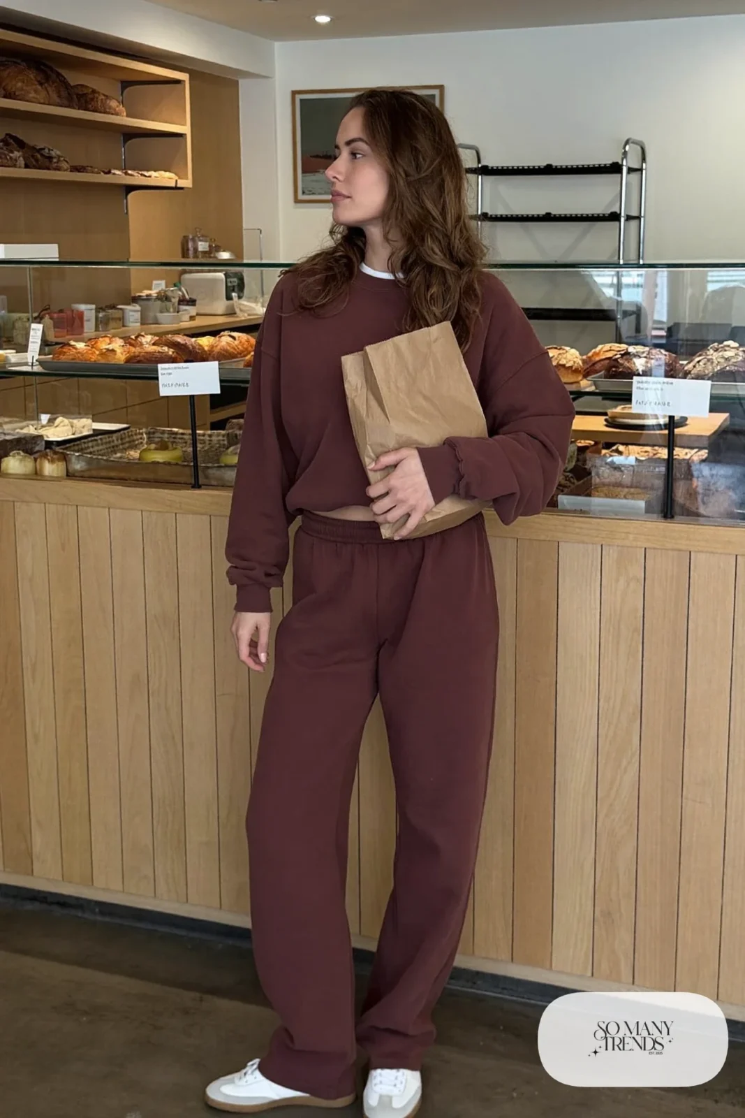 Woman in an aubergine sweatshirt and wide-leg sweatpants set with white sneakers at a bakery counter, outfit 8