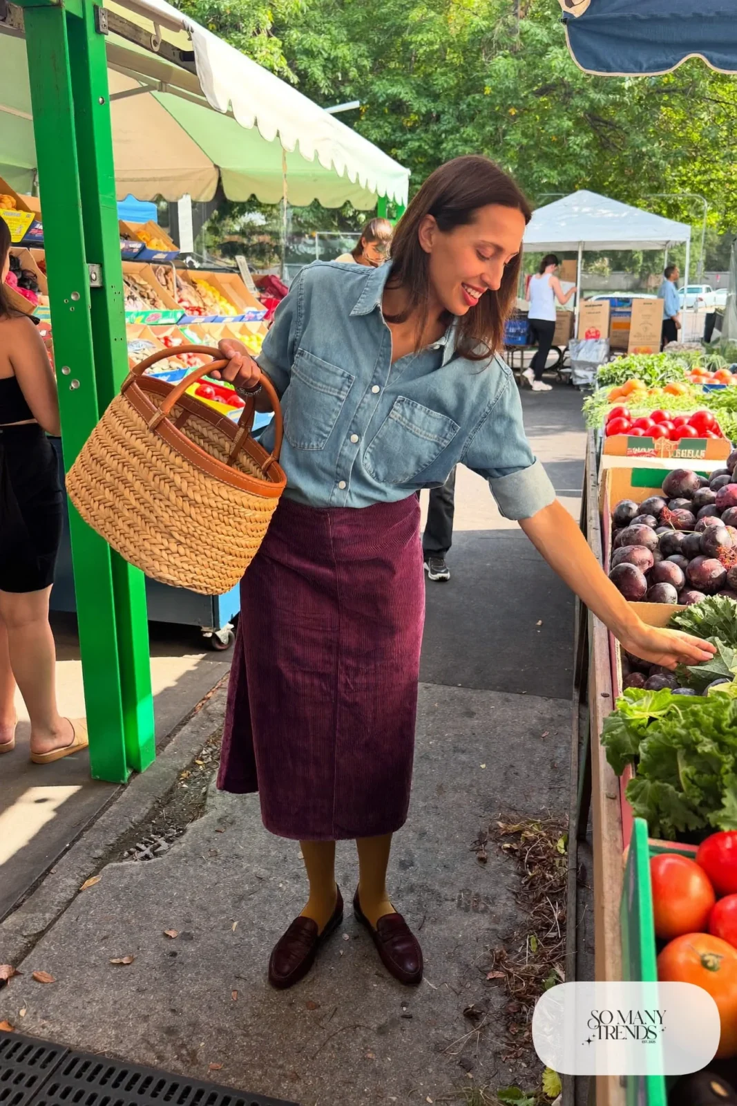Woman in a plum corduroy midi skirt, light denim shirt and brown loafers at a farmers market, outfit 10