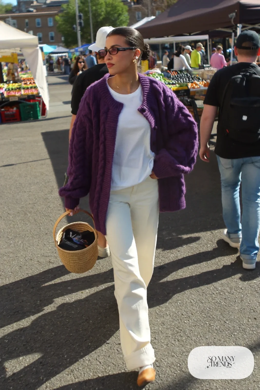 Woman in a chunky eggplant cardigan, white tee, wide cream pants and tan flat mules at a farmers market, outfit 20