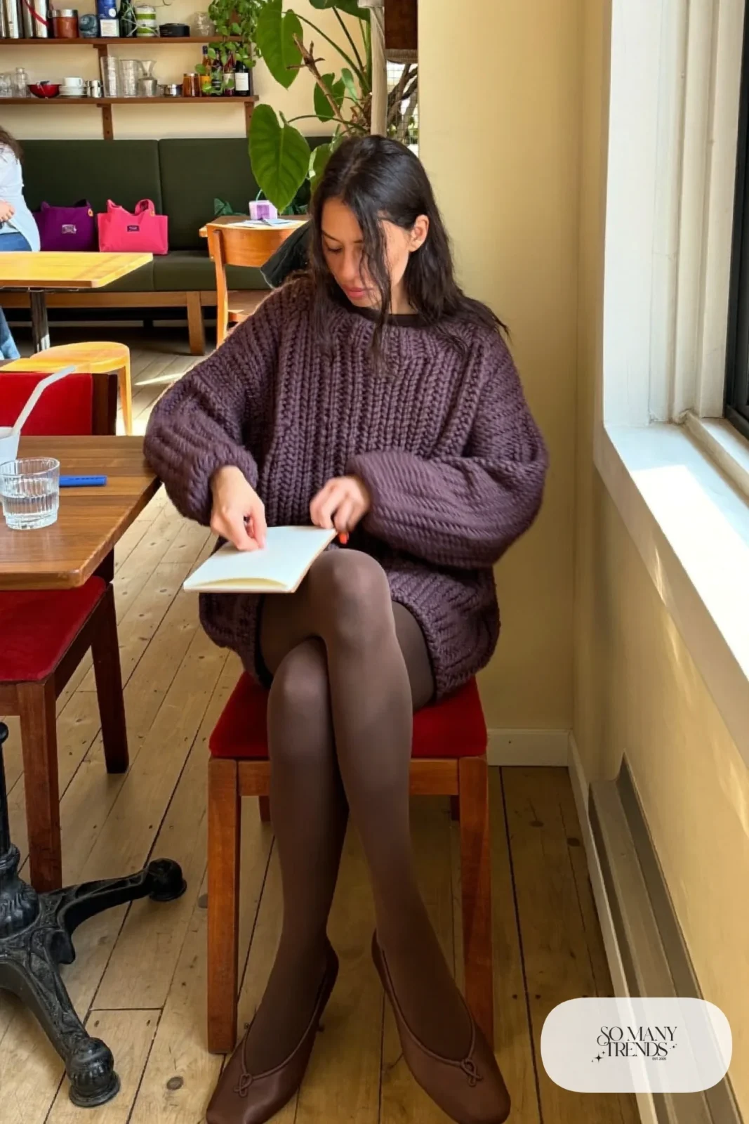 Woman in a loose aubergine sweater mini dress, thick tights and brown flats at a cozy cafe, outfit 22