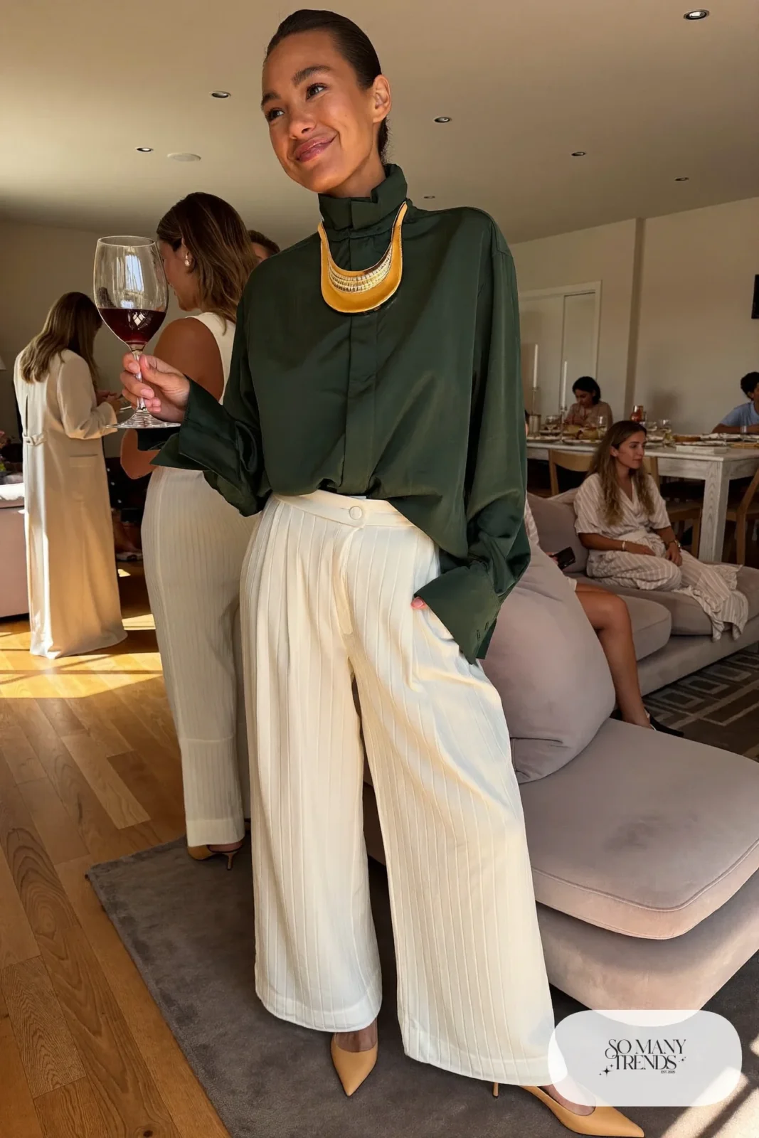 Woman in a forest green collared blouse, cream wide-leg pants, gold bib necklace and tan pumps, outfit 19