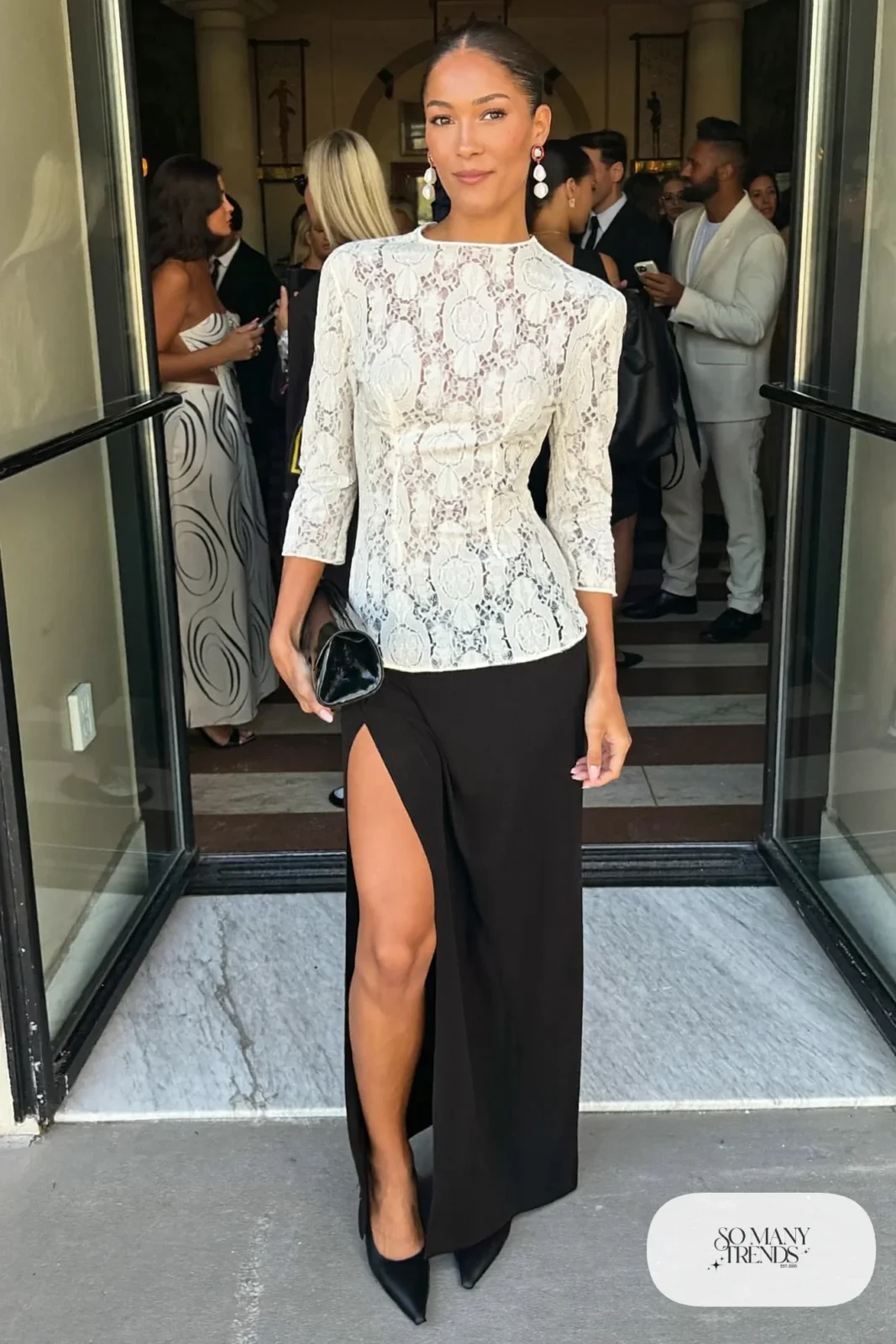 Ivory long-sleeve lace top with a long black column skirt with slit, pointed pumps and statement earrings, look 25