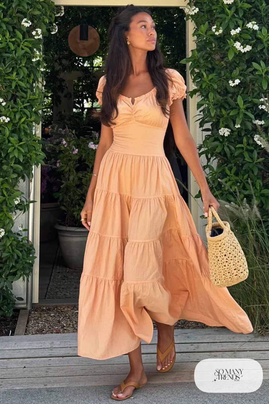 Woman in a soft peach tiered cotton maxi dress with tan flat sandals and a straw bag, garden party outfit 2