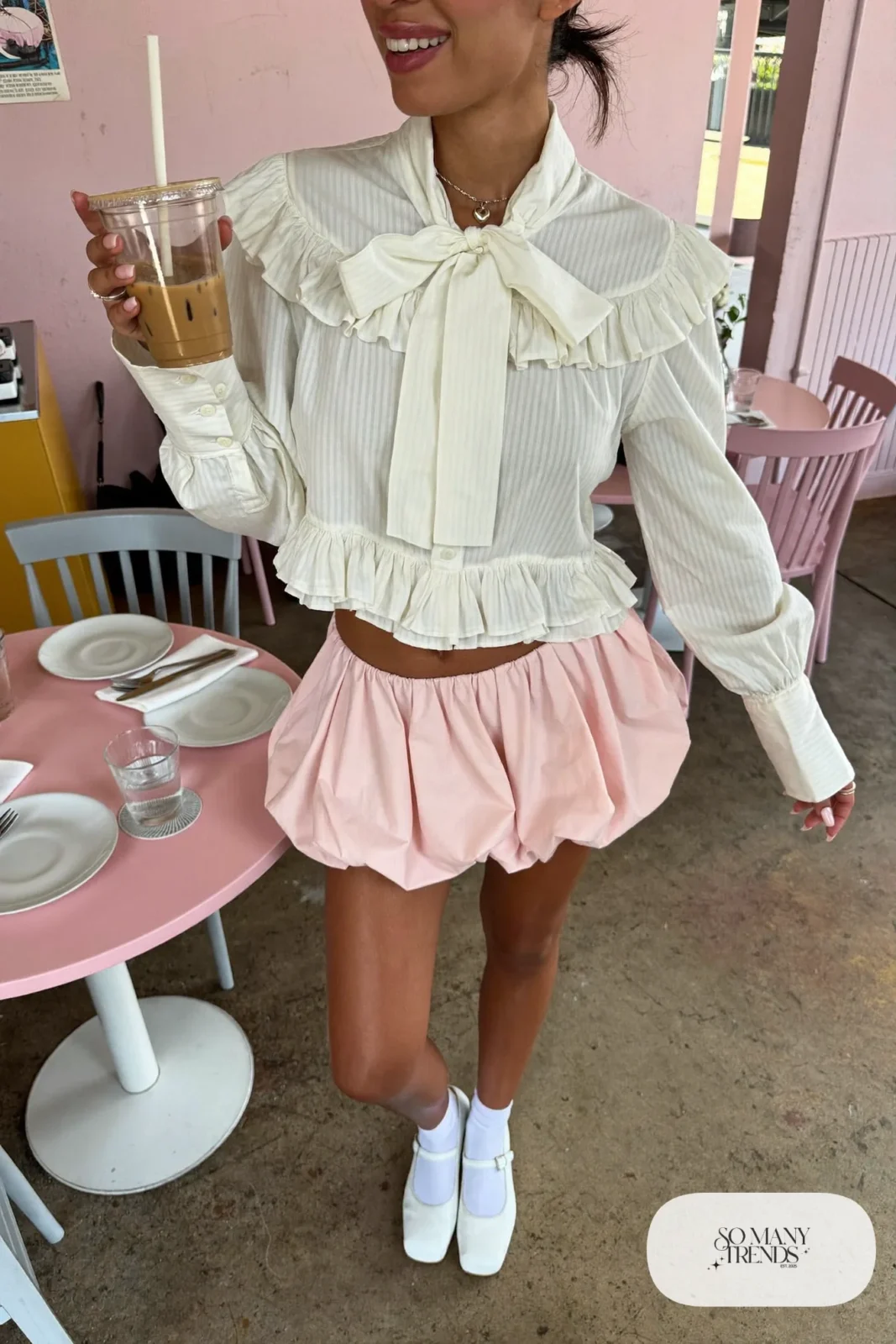 Woman in a cream ruffled bow blouse, pink bubble mini skirt and white mary janes in a pink brunch cafe, look 1