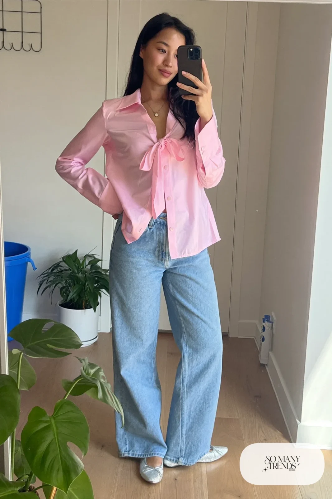 Woman in a pink bow-tie top, light relaxed jeans and sparkly flats taking a mirror selfie in a hallway, look 4