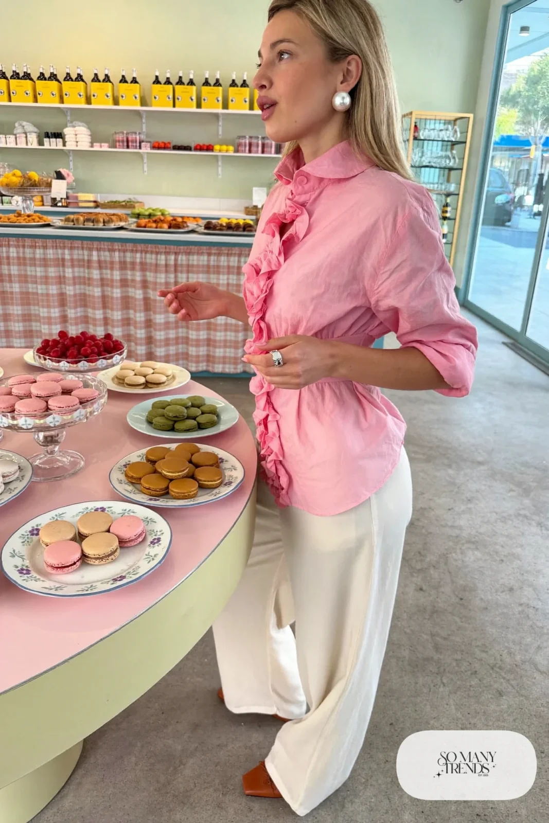 Woman in a pink ruffle collar blouse, cream wide-leg pants and cognac flats at a bakery table, look 13