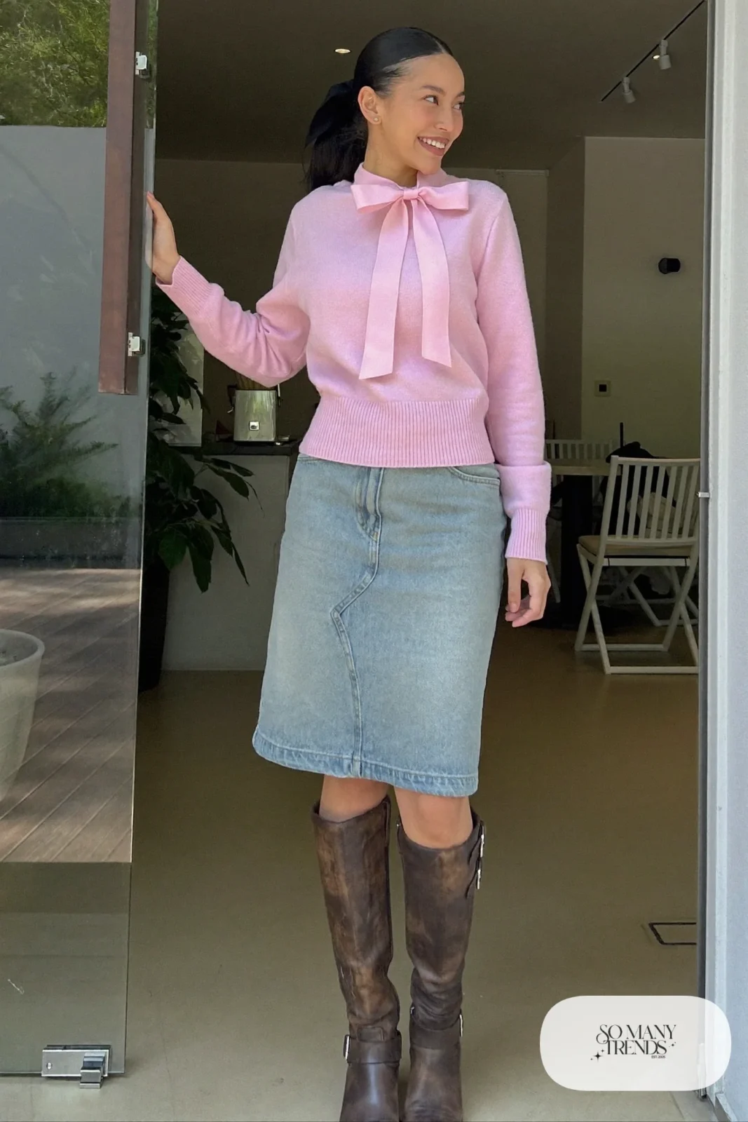 Woman in a pale pink bow sweater, denim midi skirt and brown tall boots at a cafe entrance, look 14