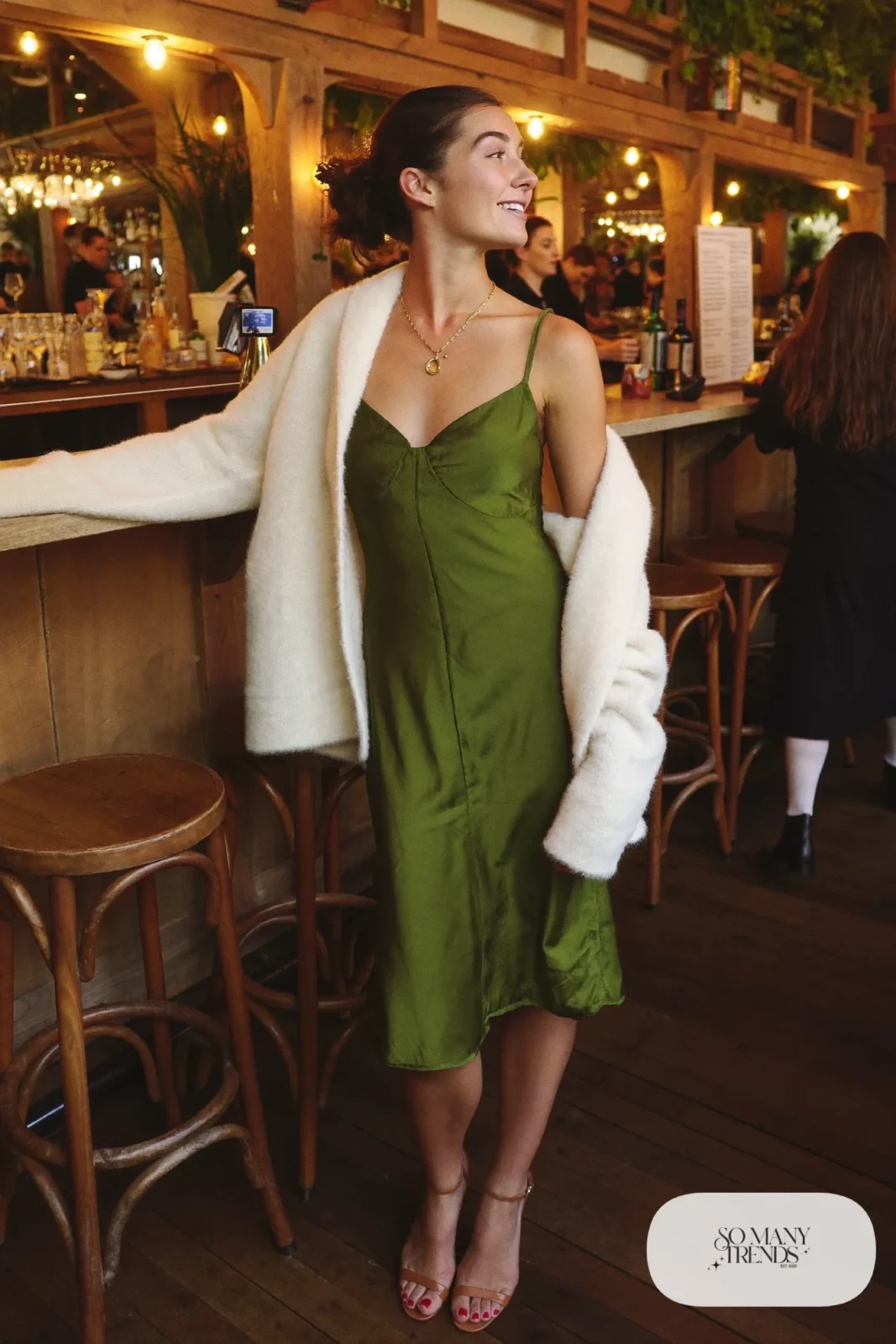 Woman in a moss green slip dress, cream fuzzy cardigan and cognac heels at a restaurant bar, look 17