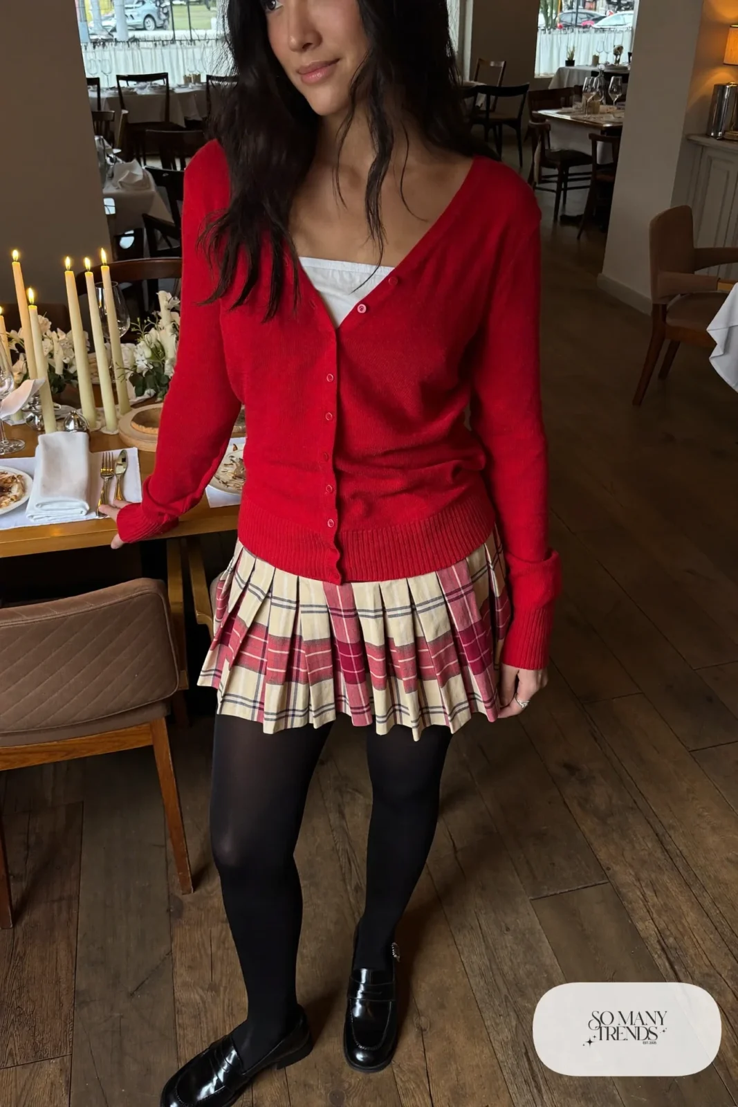 Woman in a red cardigan, white cami, plaid mini skirt, black tights and loafers at a dinner table, look 19