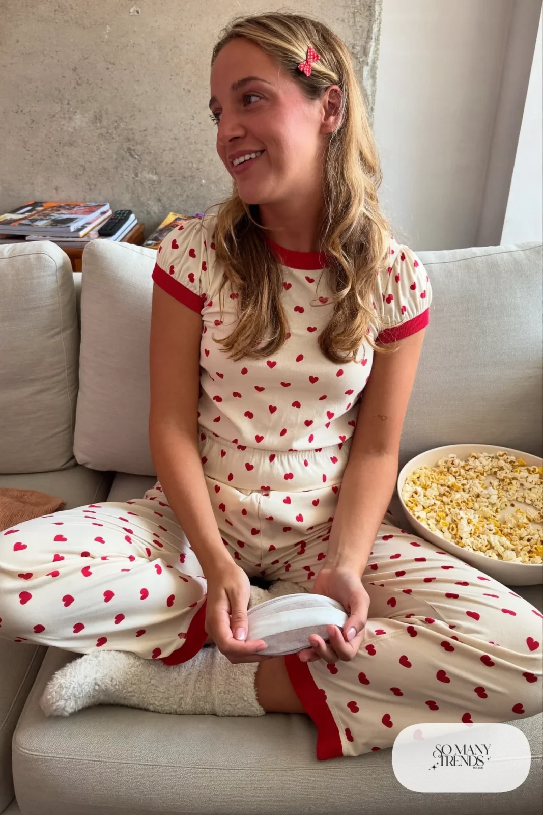 Woman in heart print cotton pajamas, fuzzy socks and a hair bow on a living room couch, look 23