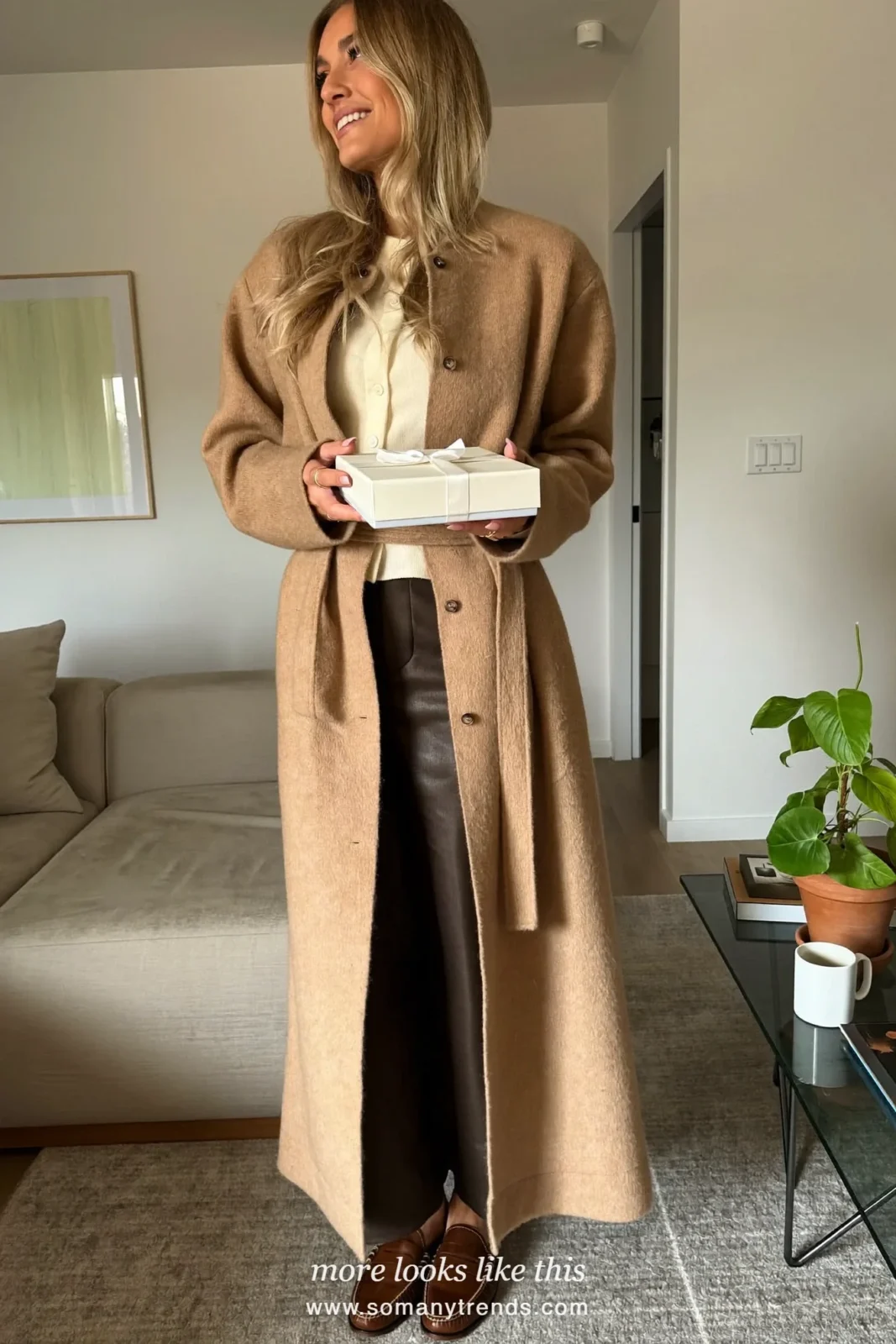 Woman in a long belted camel wool cardigan, cream knit, dark pants and brown loafers in a living room, outfit 15
