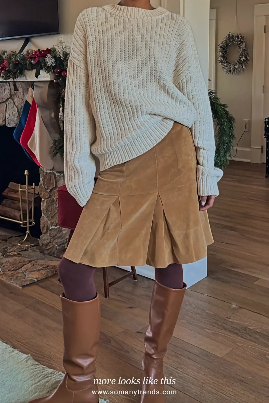 Woman in an ivory fisherman cable knit sweater, camel midi skirt and brown boots by a fireplace, outfit 5