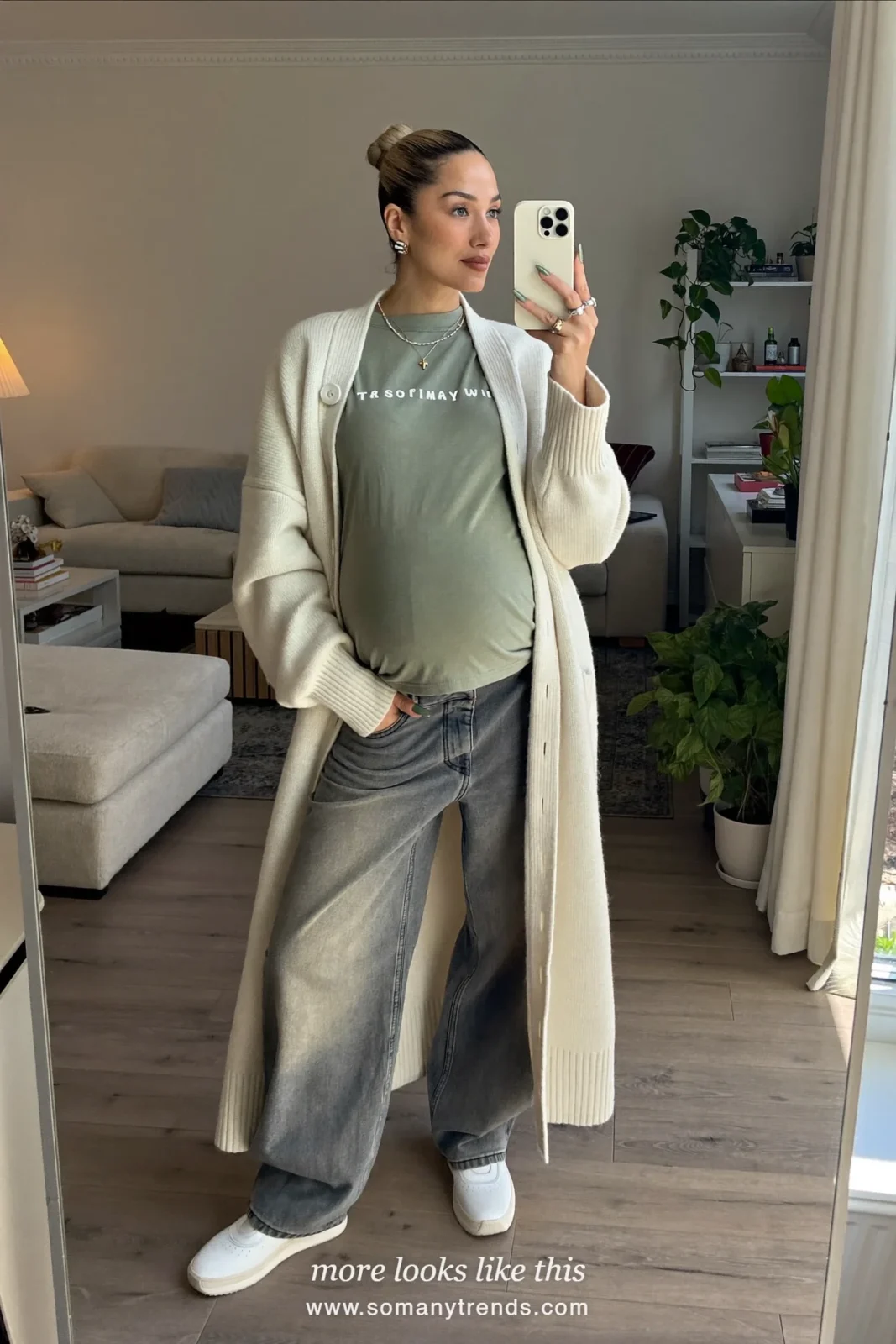 Woman with a baby bump taking a mirror selfie in a living room in an ivory cardigan, sage tee, faded maternity jeans and slip-on sneakers, outfit 12