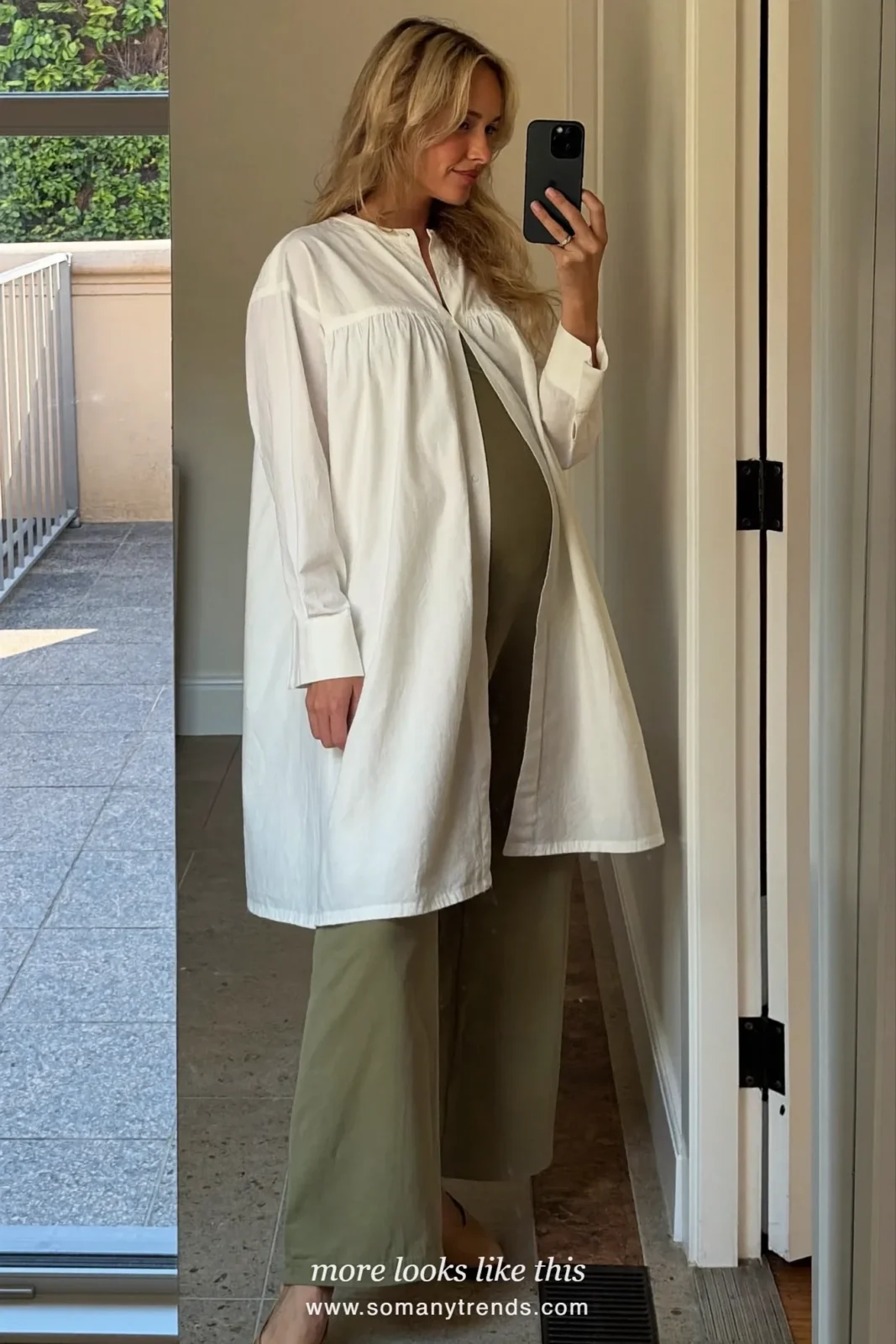 Woman with a baby bump taking a mirror selfie in an entryway in a long ivory tunic, olive maternity pants and flats, outfit 16