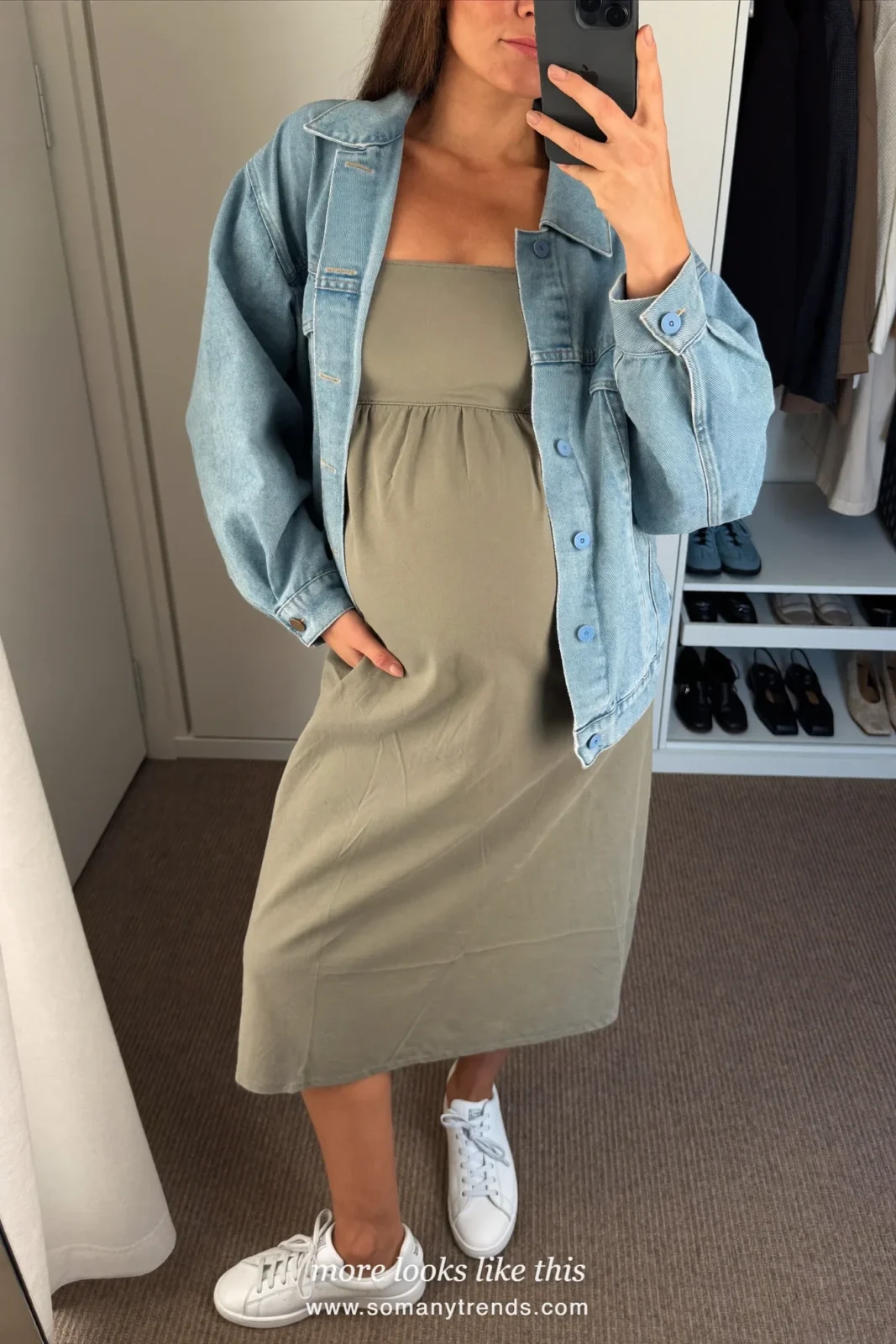 Woman with a baby bump taking a mirror selfie in a closet room in a sage midi dress, an open boxy denim jacket and sneakers, outfit 24