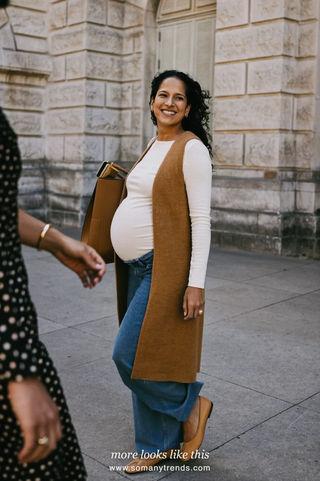 Woman with a baby bump in a long caramel knit vest over an ivory tee, wide-leg blue maternity pants and leather flats, outfit 19