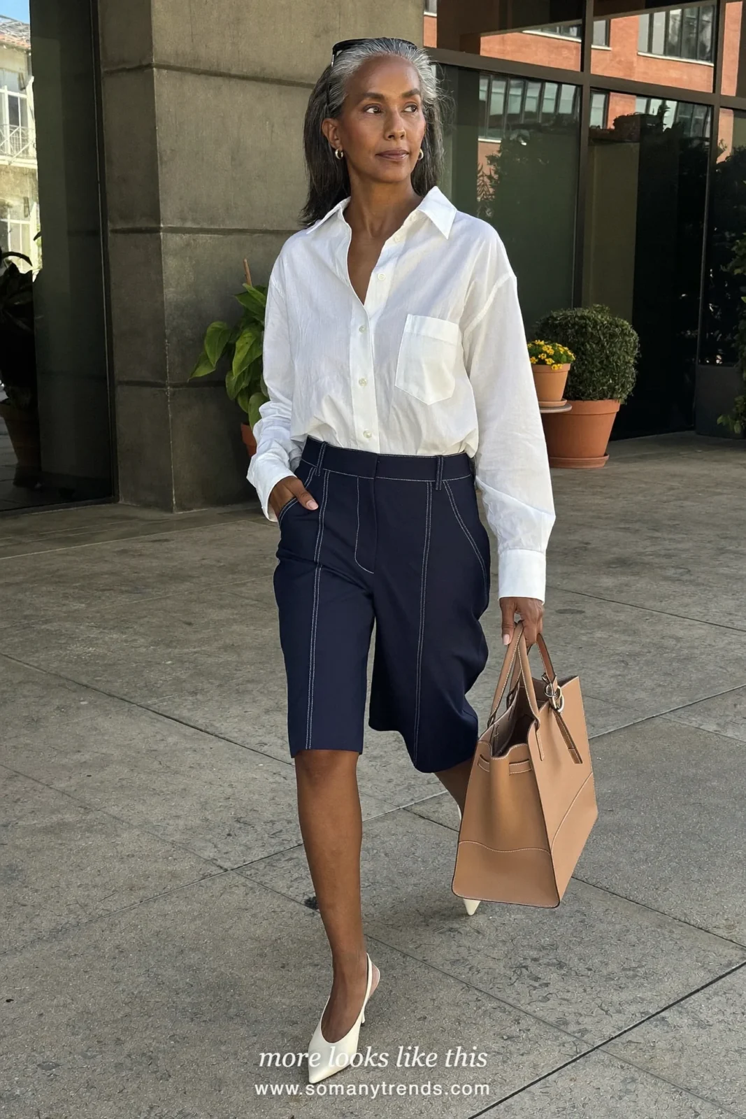 Woman in her forties wearing navy tailored Bermuda shorts, loose white poplin shirt and cream pointed pumps, outfit 1