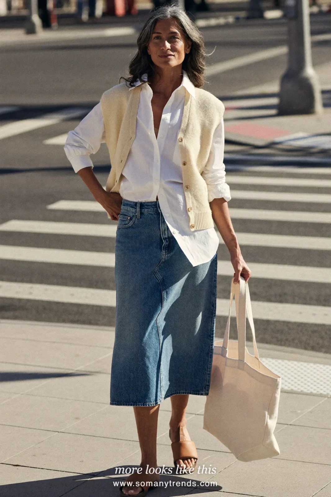 Woman in her forties wearing cream sleeveless knit vest over a white shirt, mid-wash denim midi skirt and tan leather slides, outfit 2