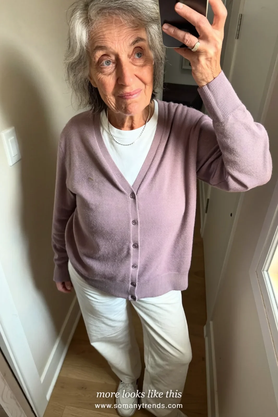 Woman in her seventies in a dusty lilac cardigan, white tee, ivory pants and white sneakers taking a mirror selfie in a hallway, look 4