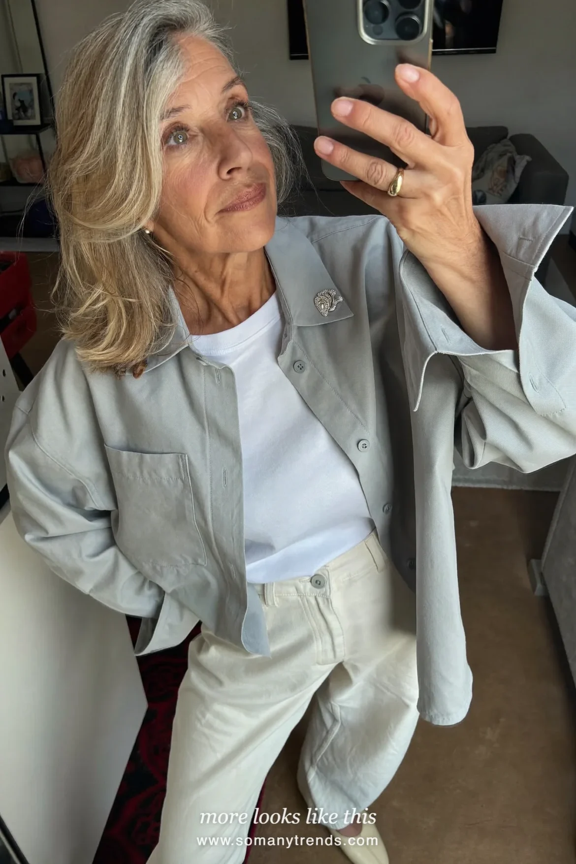 Woman in her seventies in a gray cotton shirt, white tee, ivory pants and cream flats taking a mirror selfie in a living room, look 12