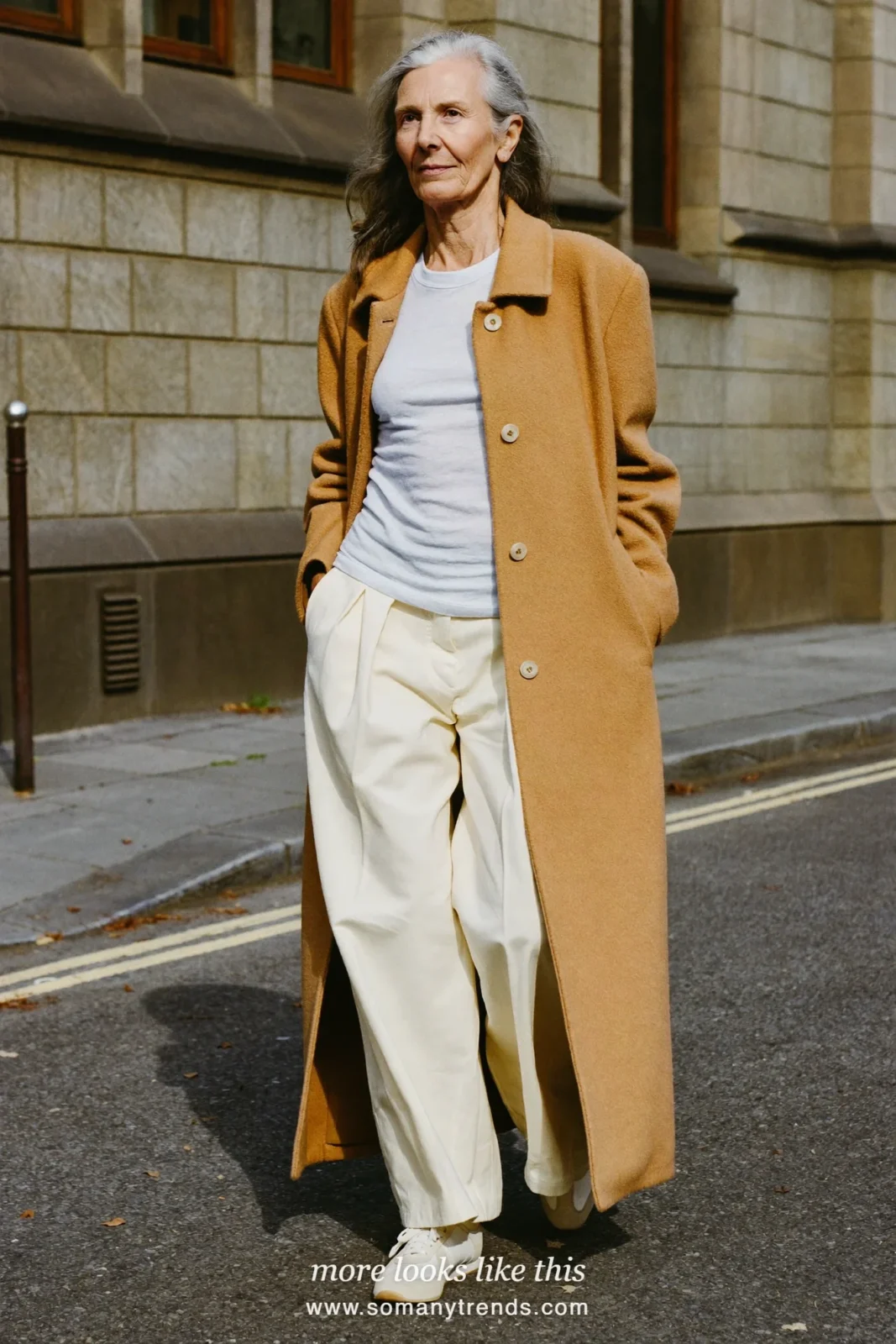 Woman in her seventies in a long camel wool cardigan coat, white tee, pale pants and cream sneakers, look 23