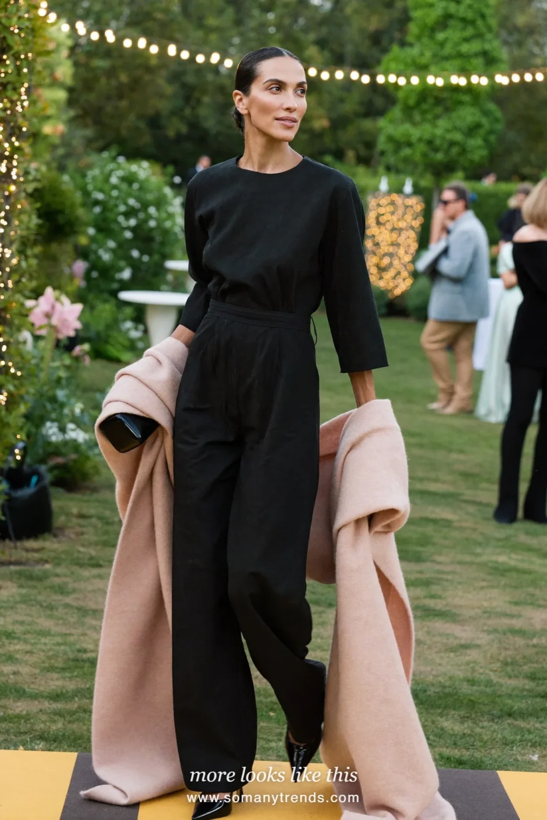 Woman in a black wide-leg crepe jumpsuit with pointed pumps and a blush wrap at a garden reception, wedding guest outfit 7