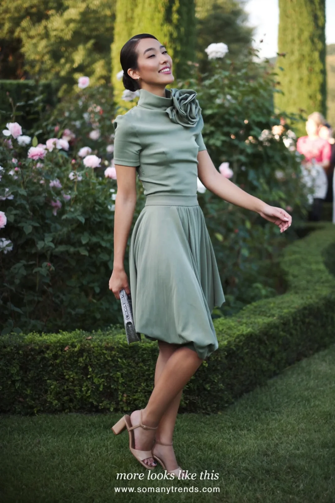 Woman in a sage green top with flower appliqué and matching midi skirt on a garden path, wedding guest outfit 17