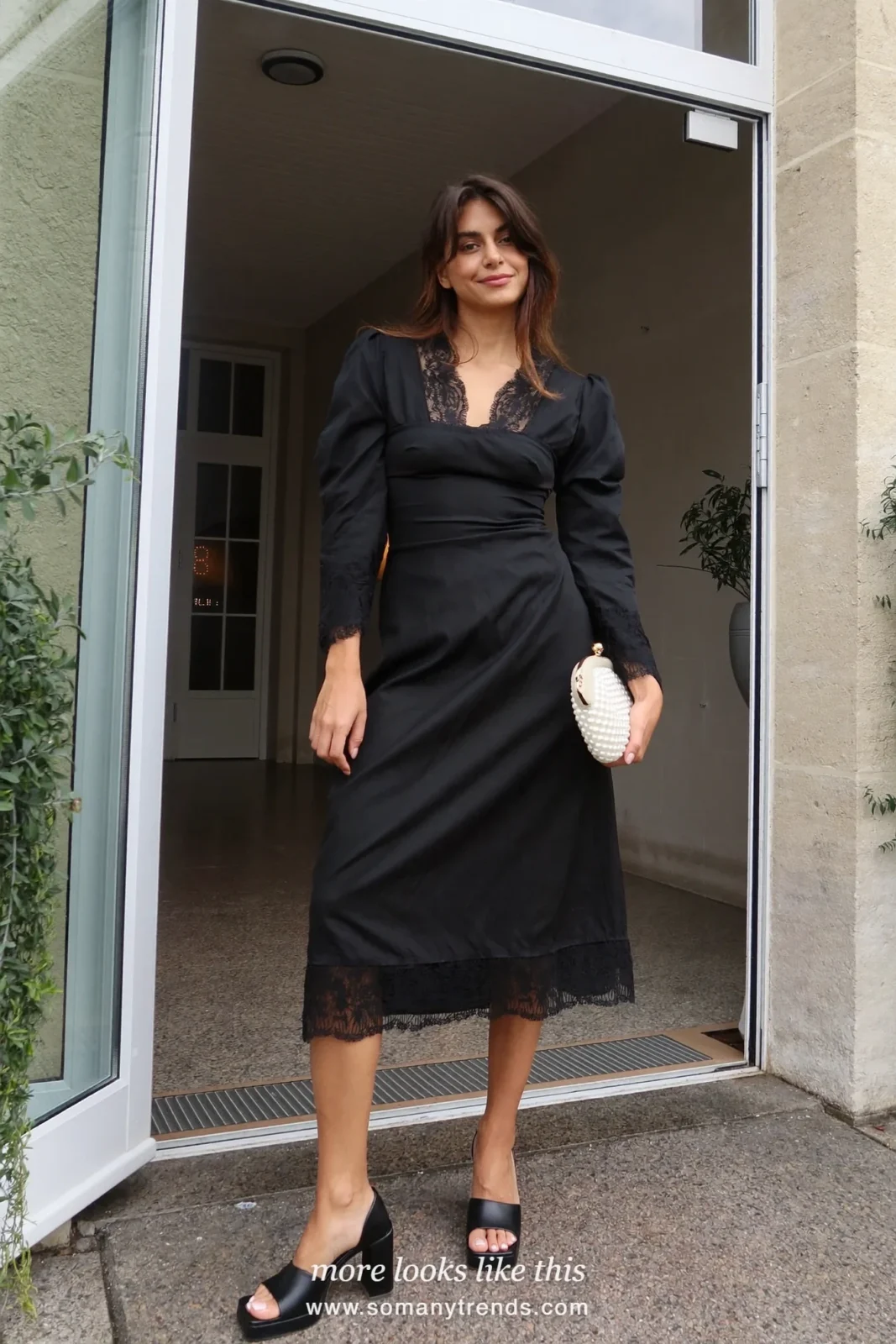 Woman in a black lace-trim midi dress with pearl clutch and block heels at a venue entrance, wedding guest outfit 13