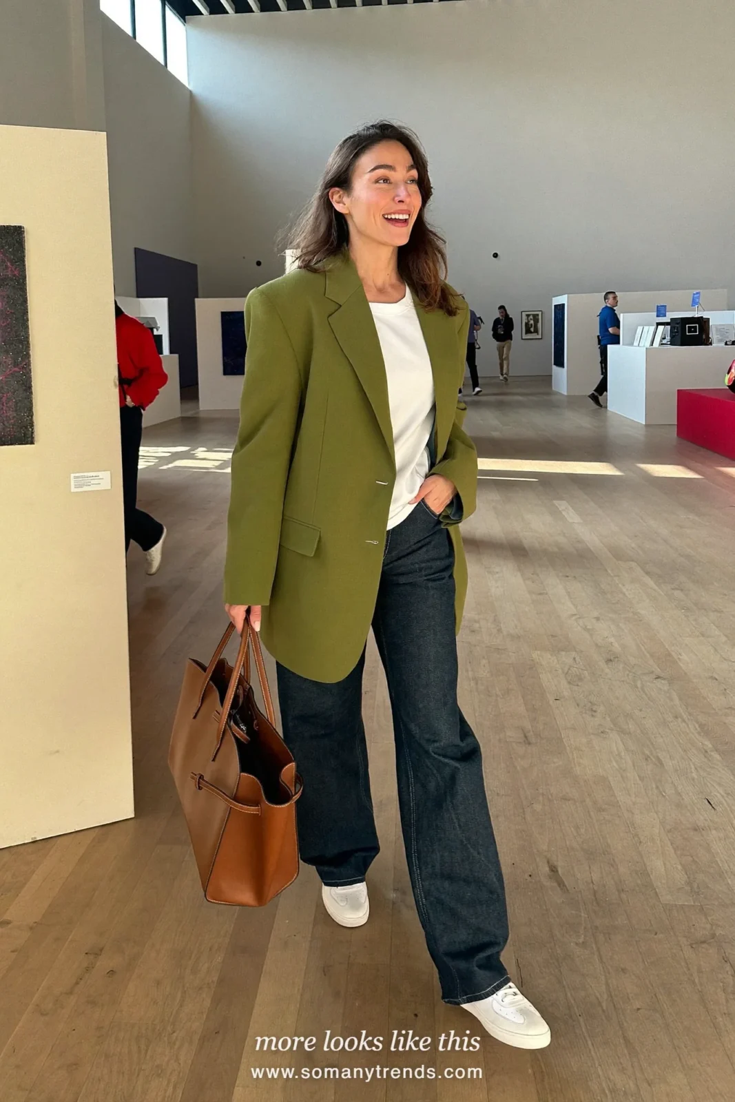 Woman in an olive oversized blazer, white tee, dark straight jeans and white sneakers on an expo floor, work conference outfit 14