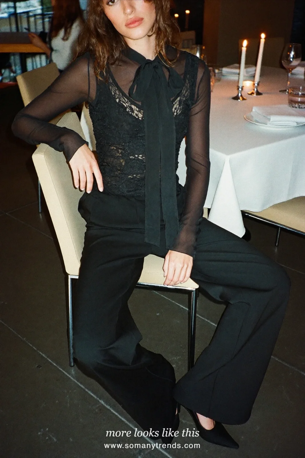Woman in a sheer black blouse with a bow, lace camisole, black wide-leg pants and pointed heels at a candlelit table, outfit 15