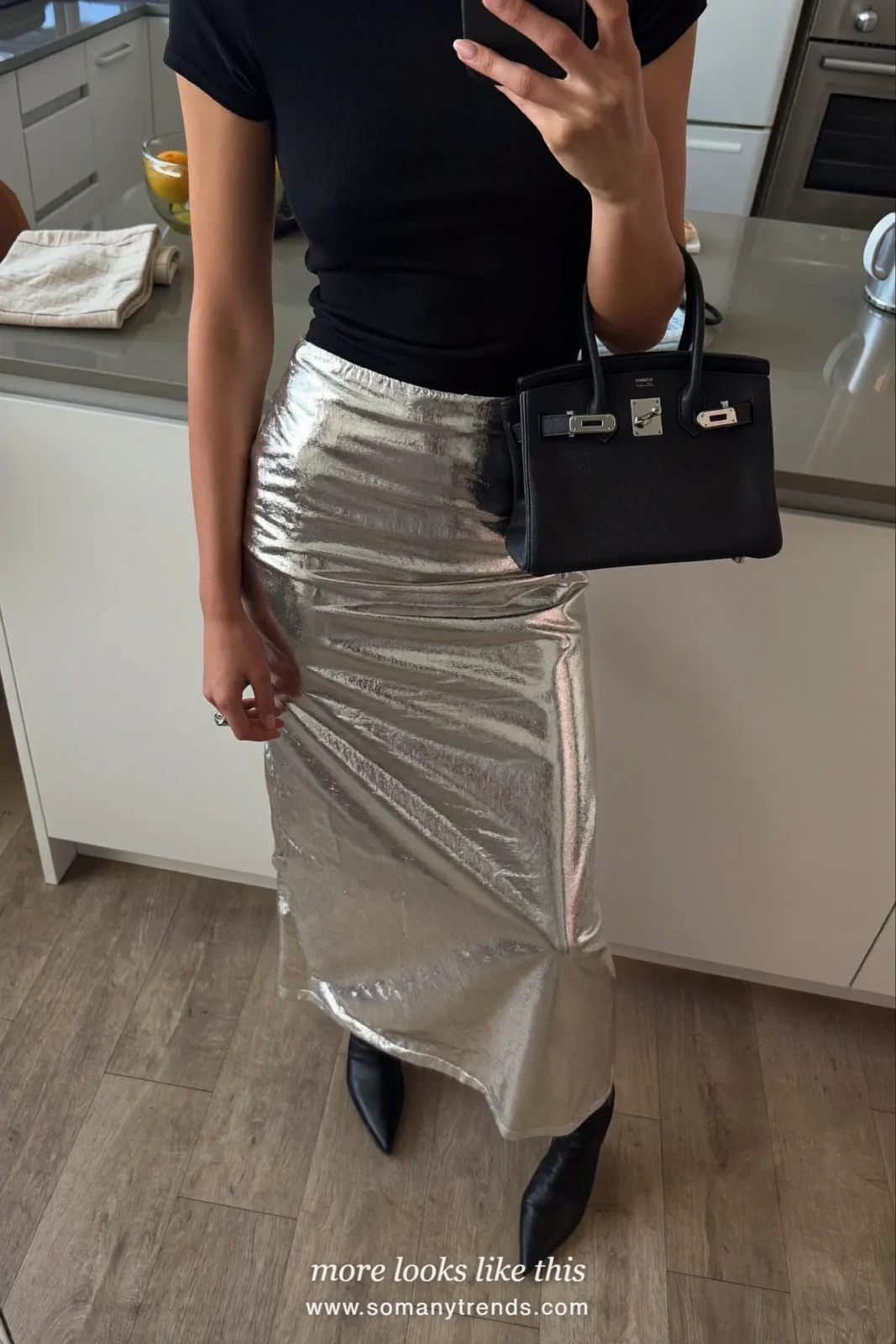 Woman in a silver metallic midi skirt, black knit top, pointed boots and a small bag outside a bar at night, outfit 20