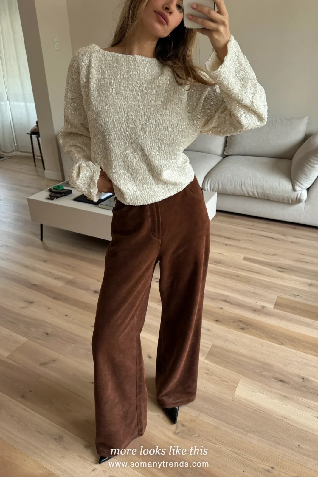 Woman in an ivory sequin top, chocolate velvet wide-leg pants and black heels taking a mirror selfie in the living room, look 4
