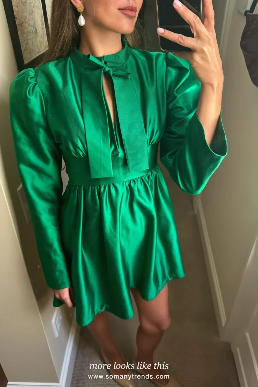 Woman in an emerald satin mini dress with a bow neckline, pearl earrings and pointed pumps taking a mirror selfie at home, outfit 4