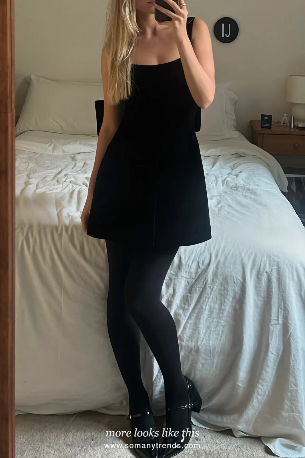 Woman in a black velvet mini dress with a bow, sheer tights and heeled mary janes taking a mirror selfie, outfit 12