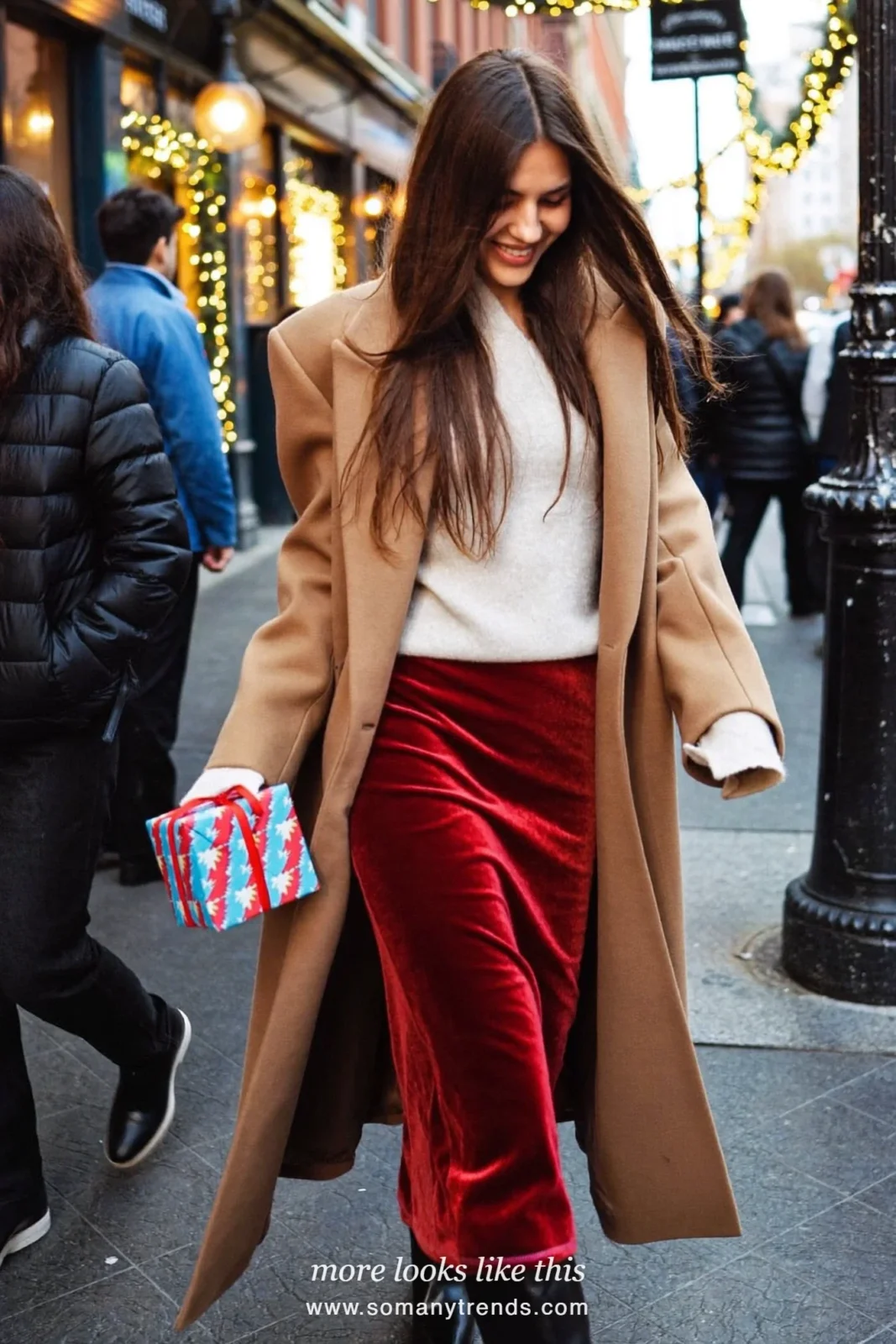Woman in a red velvet midi skirt, ivory knit sweater and tall black boots on a street with holiday lights, outfit 14